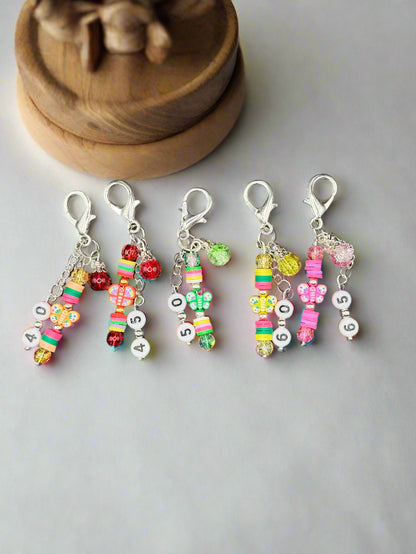Five colorful stitch markers with beads and charms on a gray background
